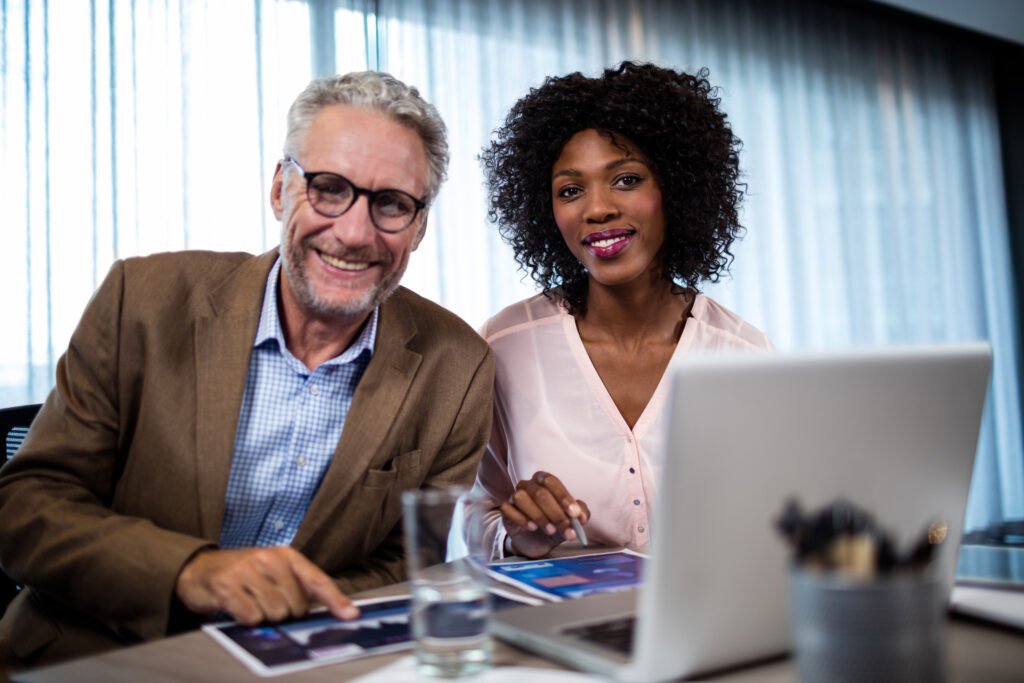 Two professionals smiling at a desk beside a laptop, meeting together as mentor and mentee.